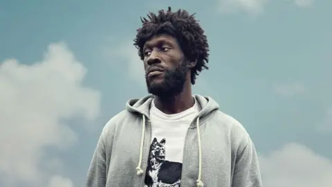 Jason Nocito Stormzy as Tenzman in Big Man. He wears a grey hoodie over a white T-shirt with a print of a dog on it. He looks to the left of the camera with a despondent expression and is shot against a pale blue cloudy sky.