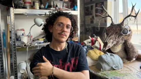 BBC A young man with glasses and dark, curly hair sits in the corner of an artist's studio. Behind him are shelves lined with materials and pots of paint and adhesives. Next to him is a workbench, where an in-progress mask resembling a deer's skull rests on top of a mannequin's head.