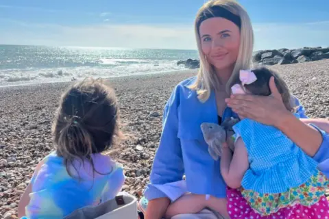 Megan Burns A woman is sat on a pebbled beach, smiling at the camera. She has a child next to her and is holding a toddler on her knee.