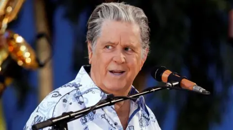 Reuters Brian Wilson appears on stage. He is wearing a white, patterned shirt and is in front of a microphone.