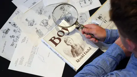 Lyon & Turnbull An aerial image showing sketches of illustrations and a copy of 'Boy' written by Roald Dahl. A person is holding a magnifying glass up to the copy of Boy. The person is wearing a blue shirt.