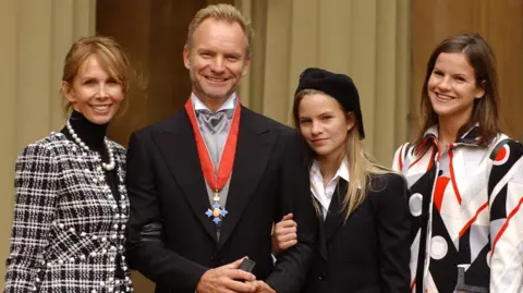 PA Sting's wife Trudie Styler in black and white checked jacket, Sting in dark suit with CBE around his neck and his daughter Coco in black jacked and Kate in white, red and black geometric shirt, at Buckingham Palace, which he received for services to the music industry. "I'm surprised and flattered to receive this honour," he said recently. "If my mum and dad were still here they would be made up."