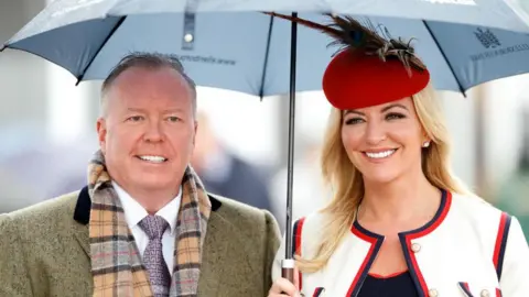 Getty Images A photograph of Doug Barrowman and Baroness Michelle Mone at the Cheltenham horse racing festival in 2019. She is carrying an umbrella and wearing a red hat and white suit. He is wearing a light grey coat and tweed scarf.