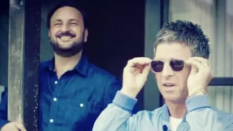Nick Brine Nick Brine and Noel Gallagher. Noel is wearing sunglasses, a blue jacket and a gold chain bracelet. Nick has dark hair and a beard and is wearing a dark blue shirt.